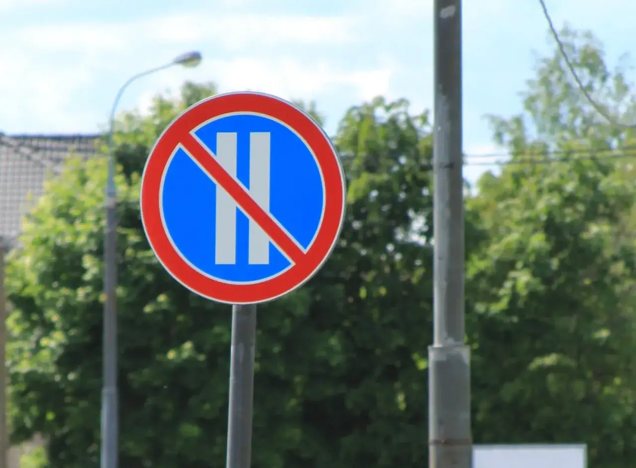 Znak B38 zakazujący postoju. Dwa pionowe białe paski na niebieskim tle, przekreślone czerwoną linią.