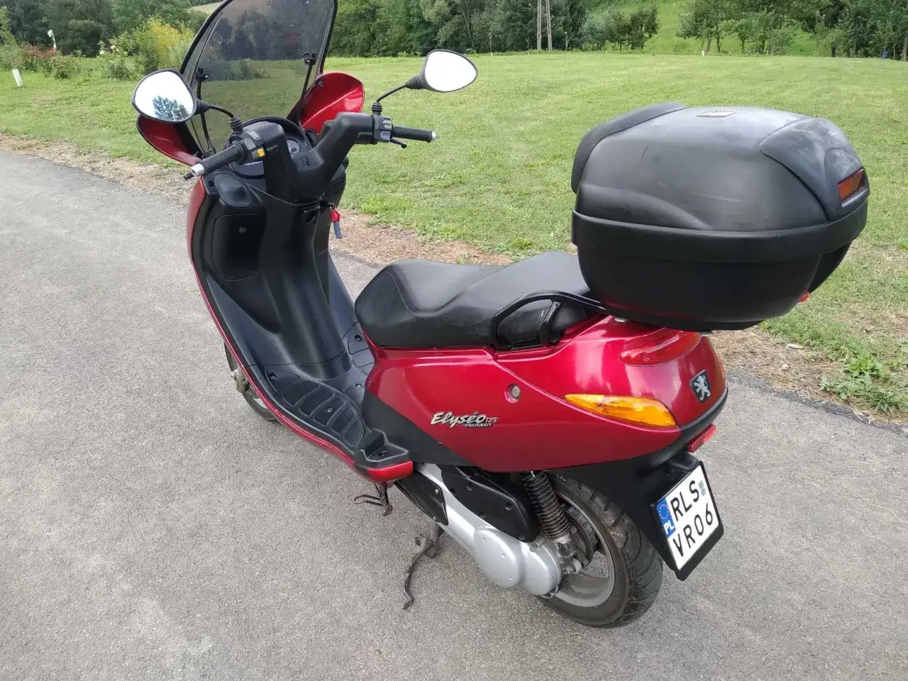 Czerwony skuter Peugeot Elyseo 125 z czarnym kufrem na bagaż, gotowy na podróż po malowniczych terenach.