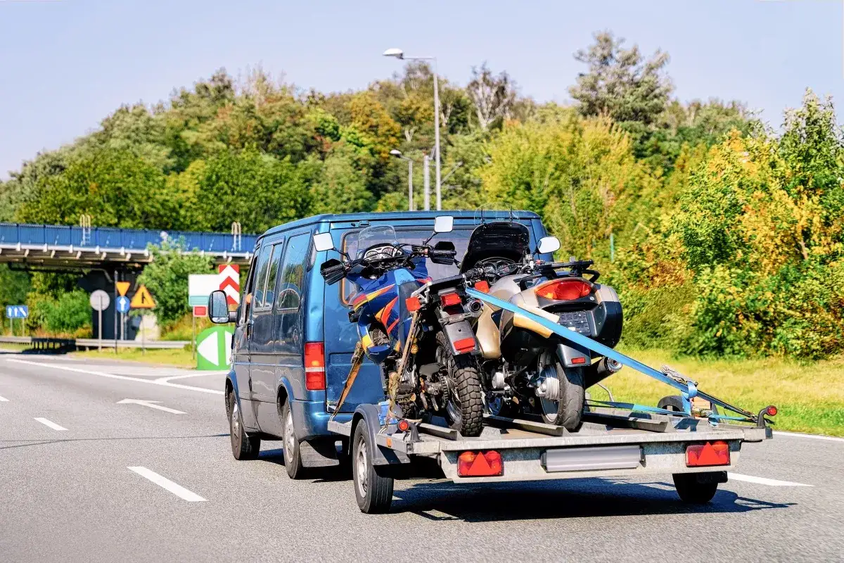 Dwa motocykle bezpiecznie przymocowane pasami na przyczepie za busem. Pokazano, jak przewieźć motocykl na zwykłej przyczepie.