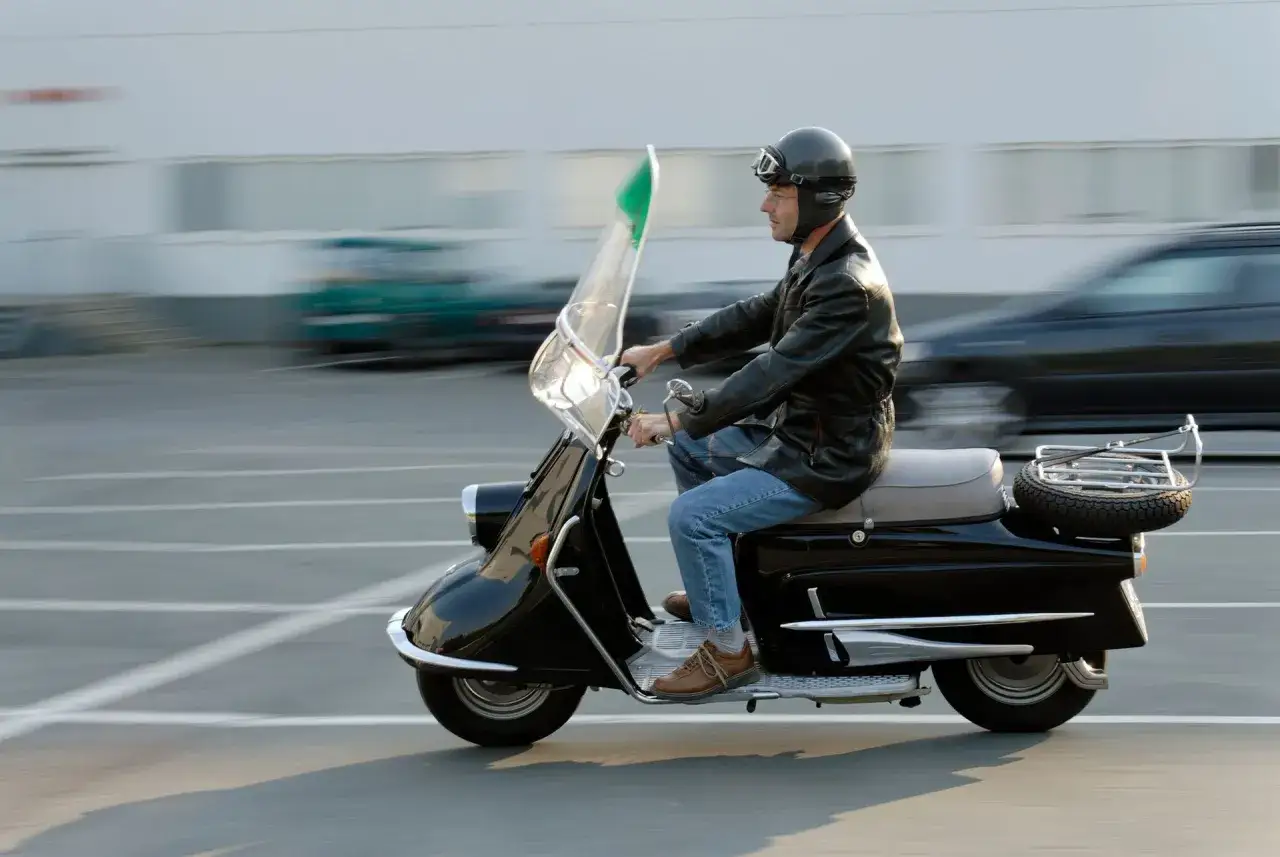 Mężczyzna w kasku i goglach jedzie czarnym motorowerem. Czy można jeździć motorowerem na dowód? Ten stylowy pojazd wygląda na taki, którym można się poruszać bez prawa jazdy.
