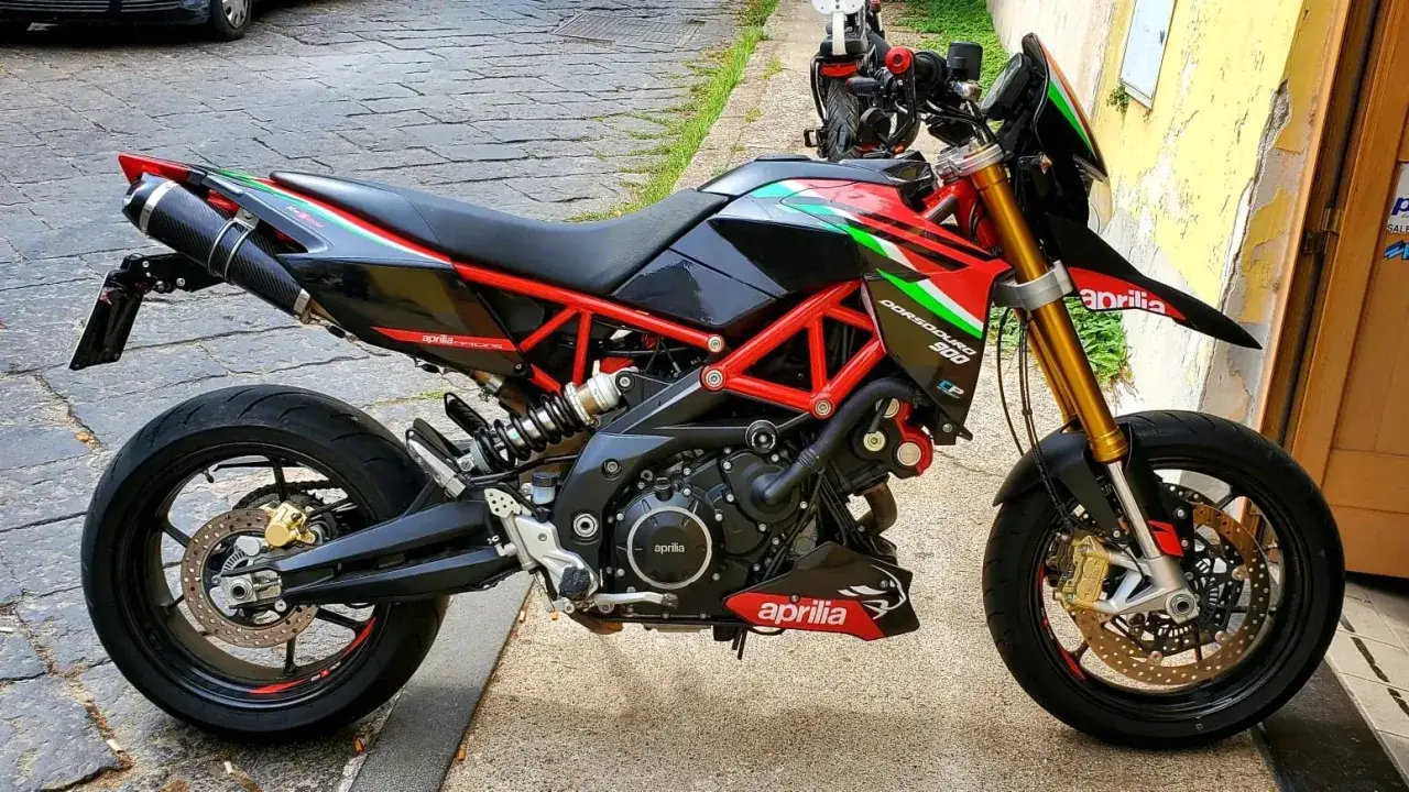 Stojący na chodniku motocykl Aprilia Dorsoduro 750 w kolorach włoskiej flagi, z czerwonym stelażem i złotym zawieszeniem.