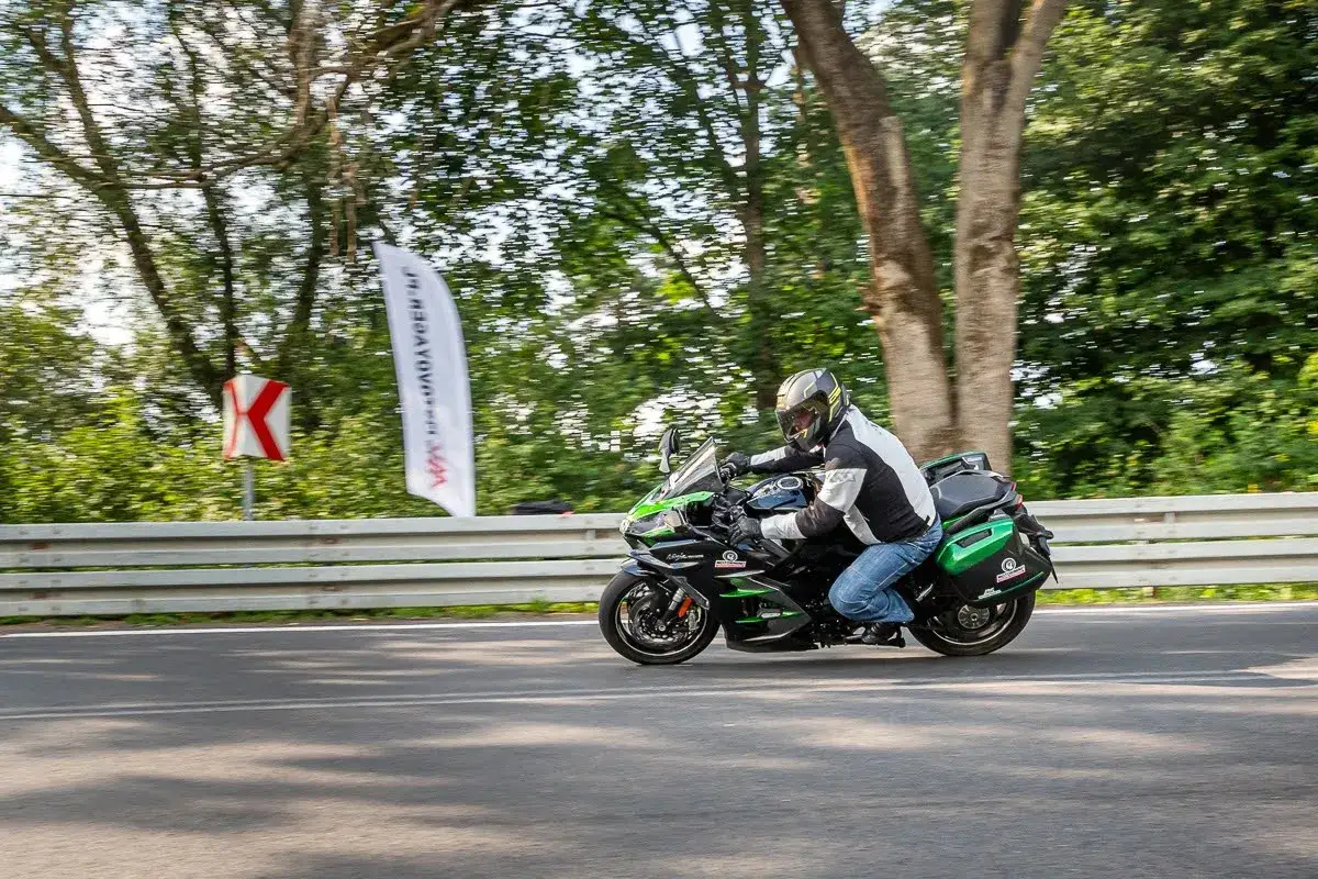 Motocyklista na czarnym Kawasaki H2 z zielonymi akcentami mknie po drodze, otoczony zielenią i drzewami.