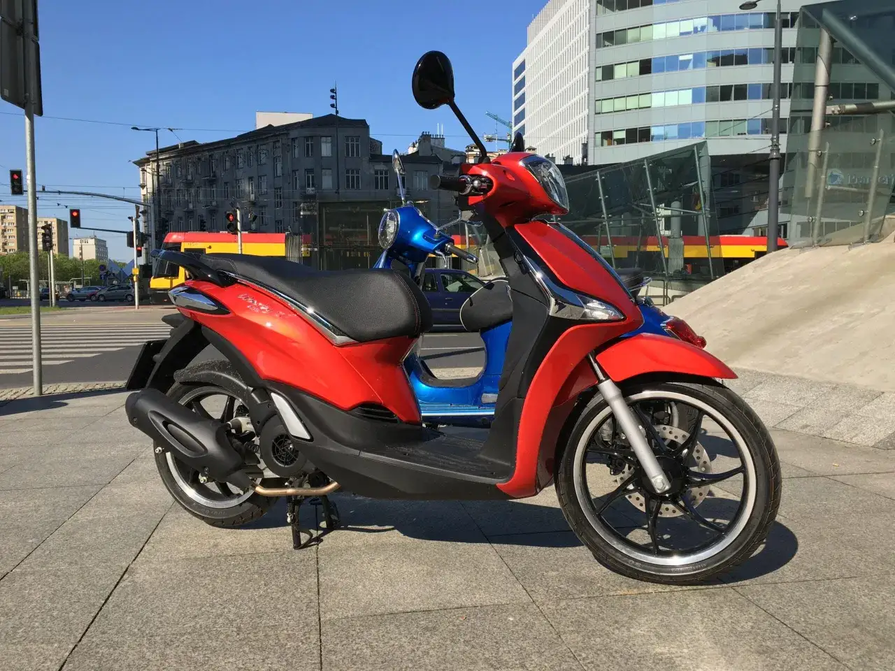 Czerwony Piaggio Liberty 125, z niebieskim skuterem w tle, gotowy do jazdy. Idealny do miasta, sprawdź piaggio liberty 125 dane techniczne.