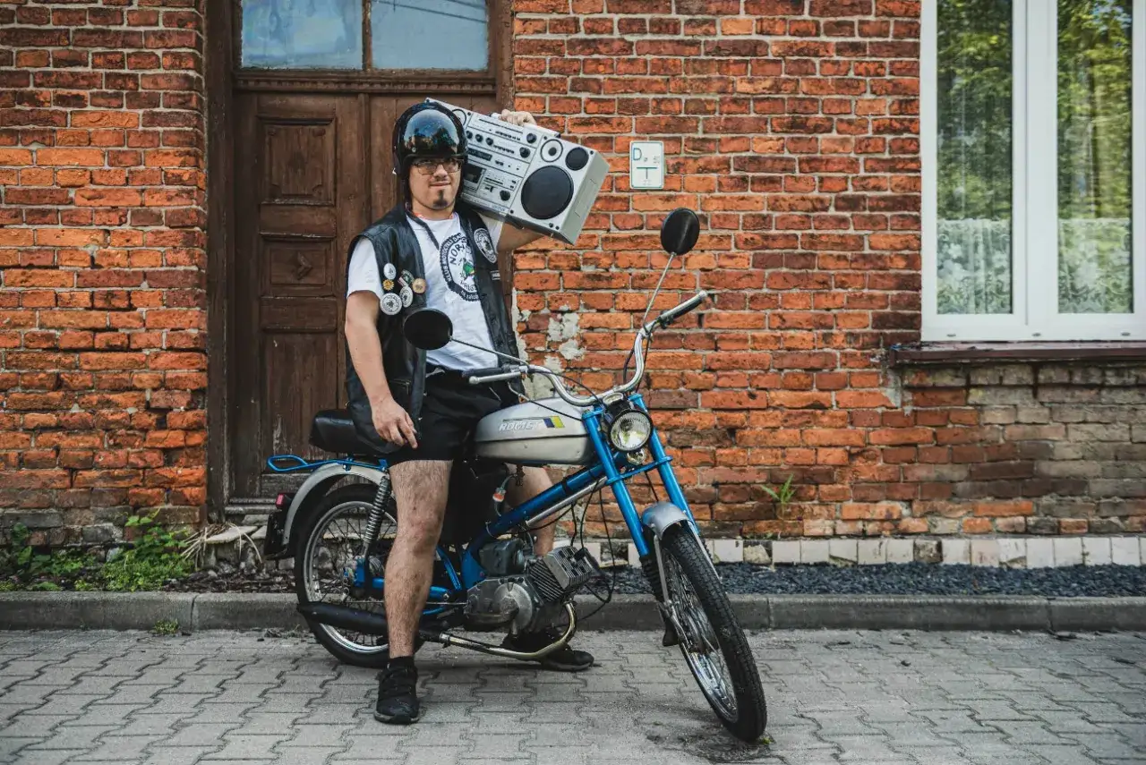 Młody mężczyzna z boomboxem na ramieniu, w kasku i kamizelce, siedzi na kultowym polskim motorowerze Romet.