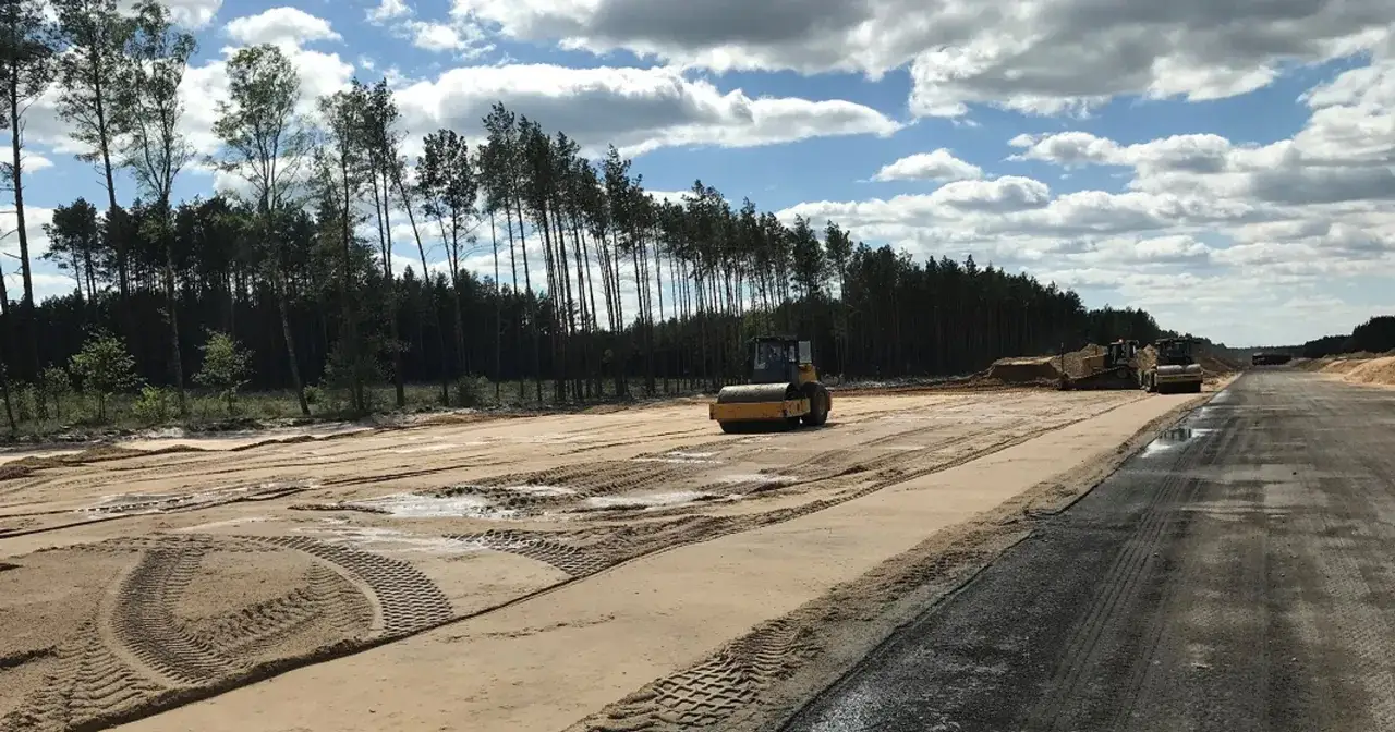Budowa drogowych odcinków lotniskowych w Polsce. Walec drogowy ubija nawierzchnię, w tle las i maszyny budowlane.