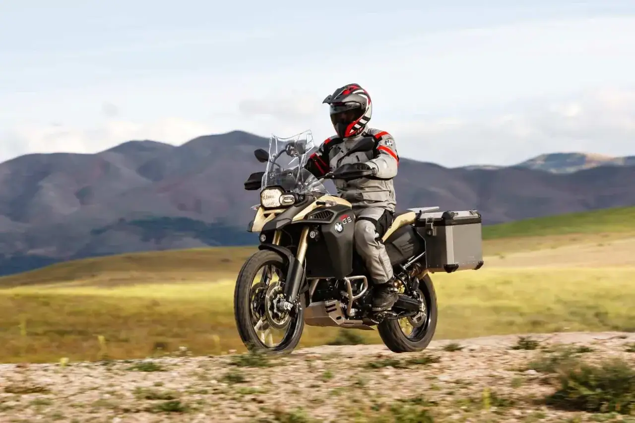 Motocyklista w kasku i stroju podróżuje na motocyklu BMW 800 GS po piaszczystej drodze, w tle górzysty krajobraz.