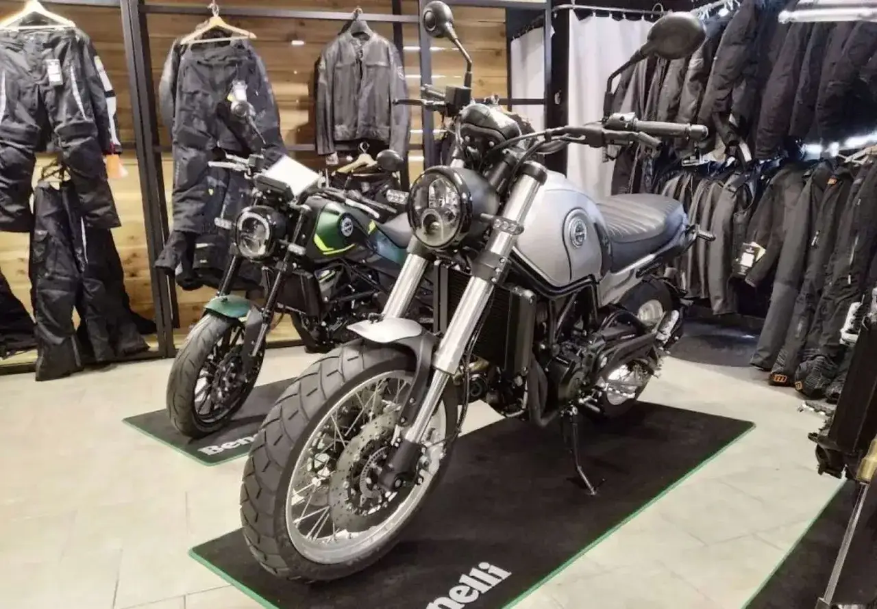 Dwa motocykle Benelli Leoncino 500 Trail w salonie, jeden srebrny, drugi zielony, otoczone odzieżą motocyklową.