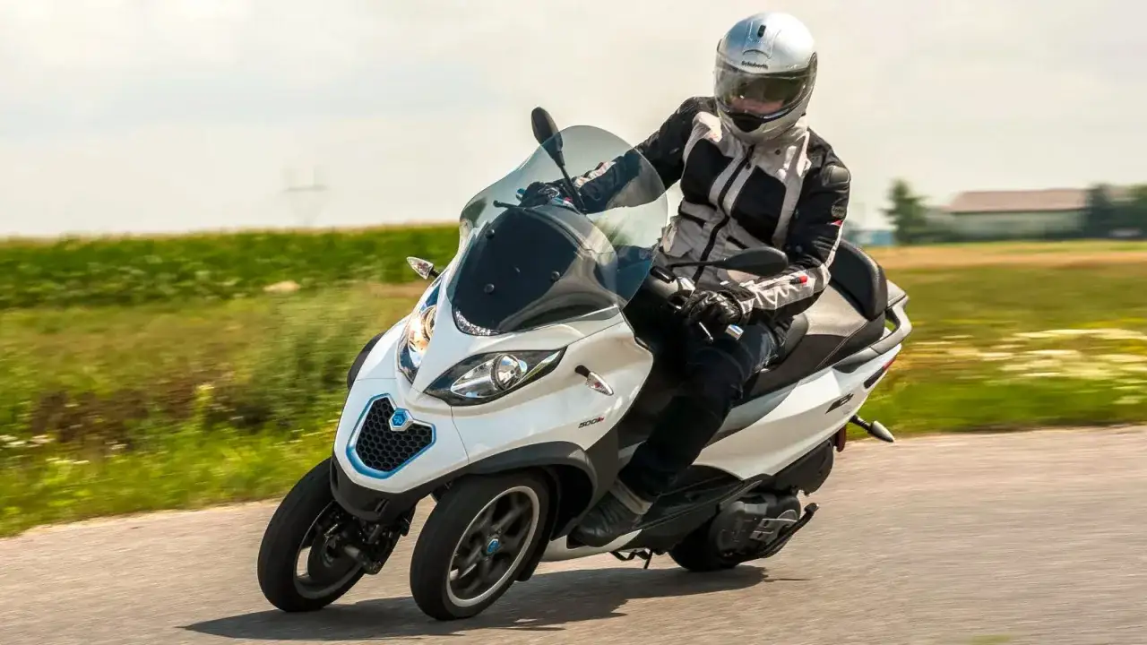 Biały Piaggio MP3 500 z kierowcą w kasku i kurtce jedzie po drodze. Poznaj piaggio mp3 500 dane techniczne.