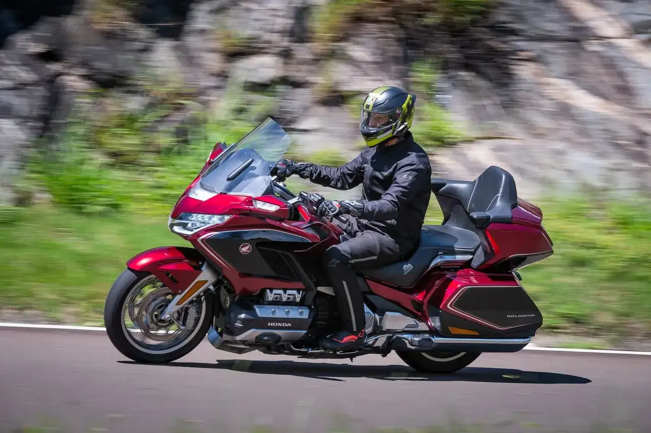 Mężczyzna w kasku jedzie lśniącym, czerwonym motocyklem honda turystyk. W tle rozmyte zielone wzgórza i skały.