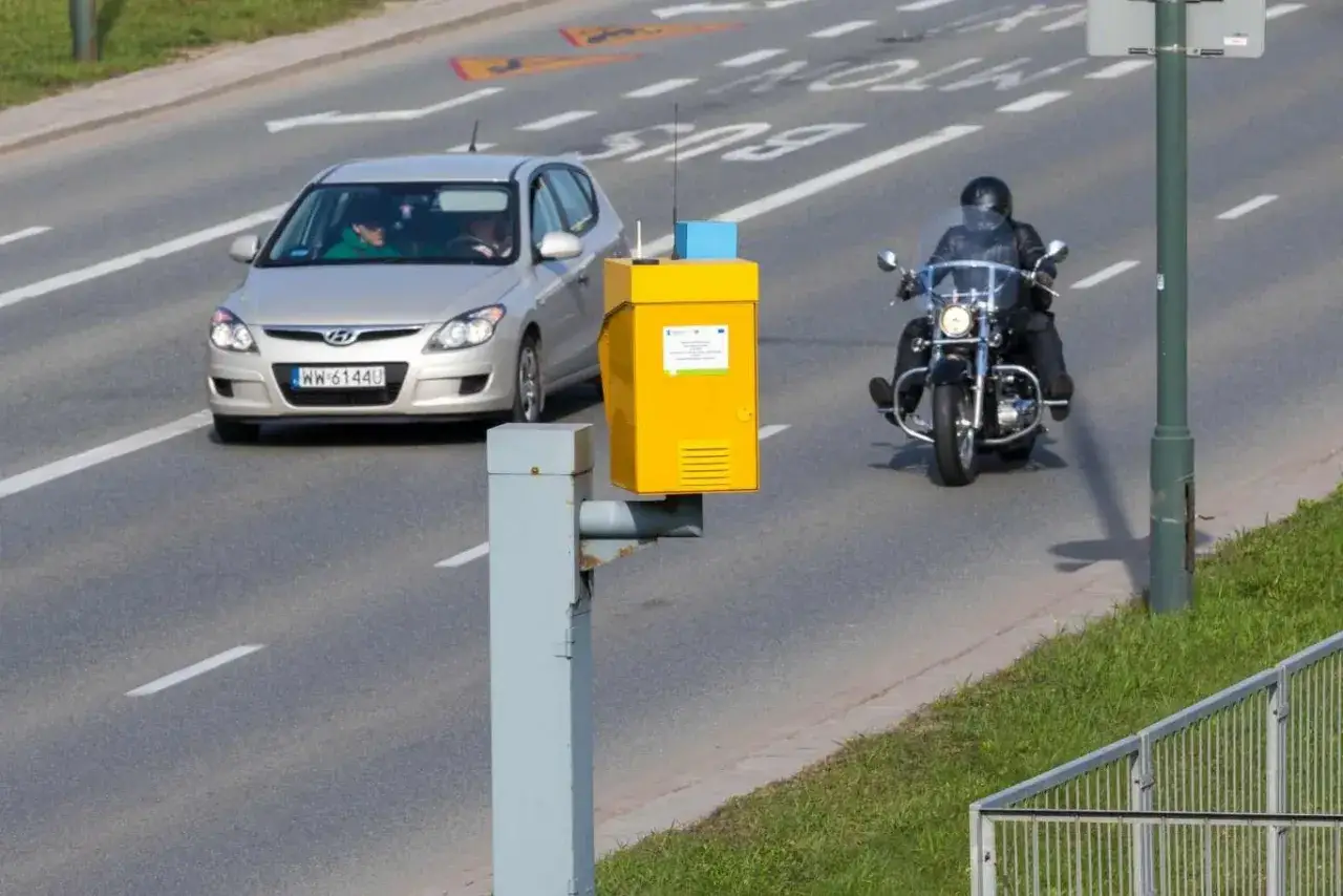 Żółty fotoradar na drodze, obok przejeżdża srebrny samochód i motocykl w czarnej kurtce.