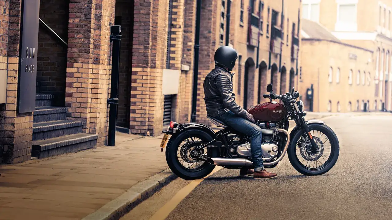 Mężczyzna w skórzanej kurtce siedzi na stylowym motocyklu Triumph Bonneville Bobber, zaparkowanym na brukowanej ulicy.