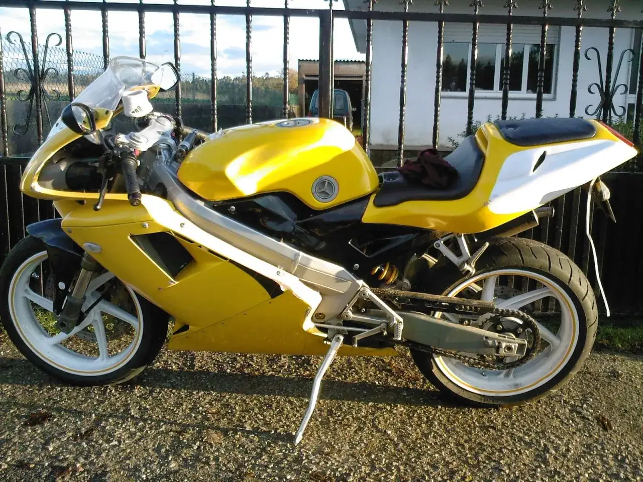 Żółta Cagiva Mito 125 V Max, gotowa do jazdy, z czarnym siedzeniem i białym zadupkiem, stoi przed ogrodzeniem.