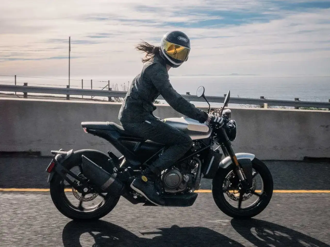 Kobieta w kasku jedzie motocyklem Husqvarna Vitpilen 701 wzdłuż wybrzeża.