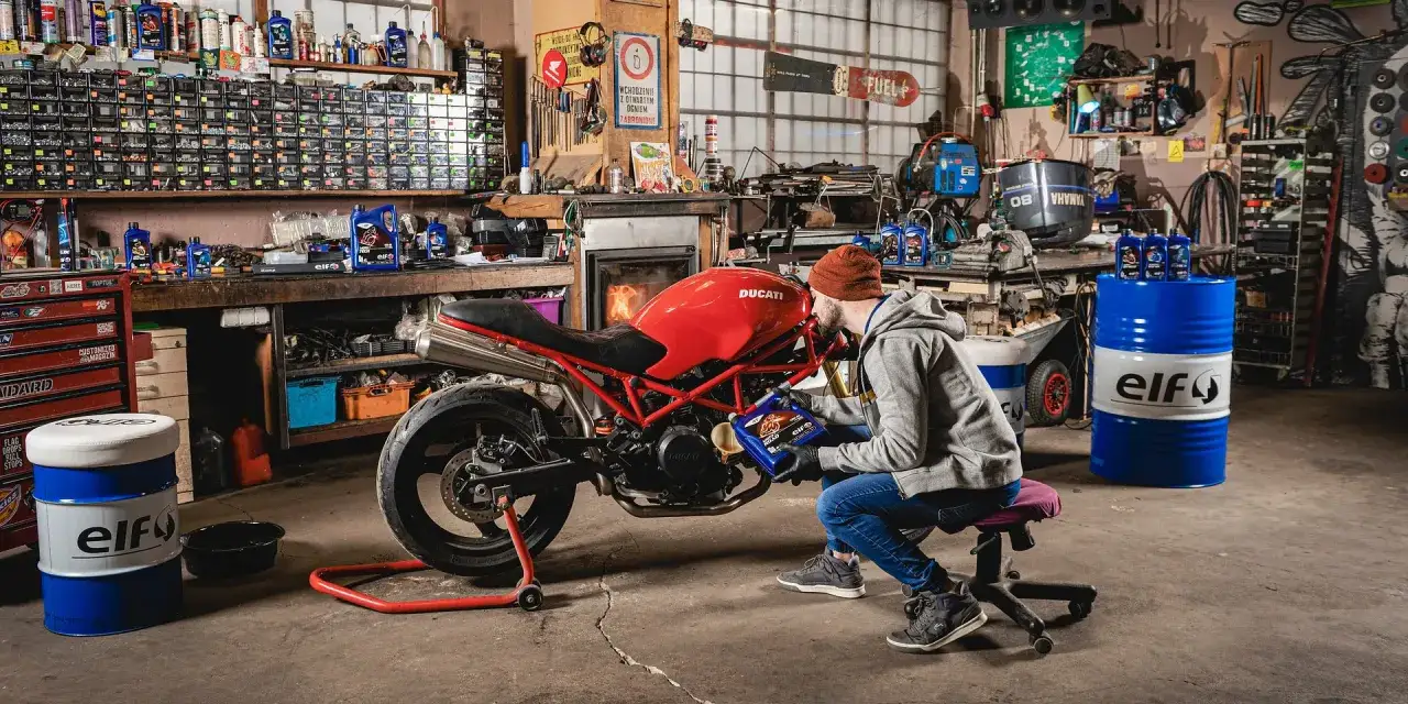 Mężczyzna w warsztacie dolewa płyn chłodniczy do czerwonego Ducati. W tle półki z narzędziami i beczki z logo Elf.