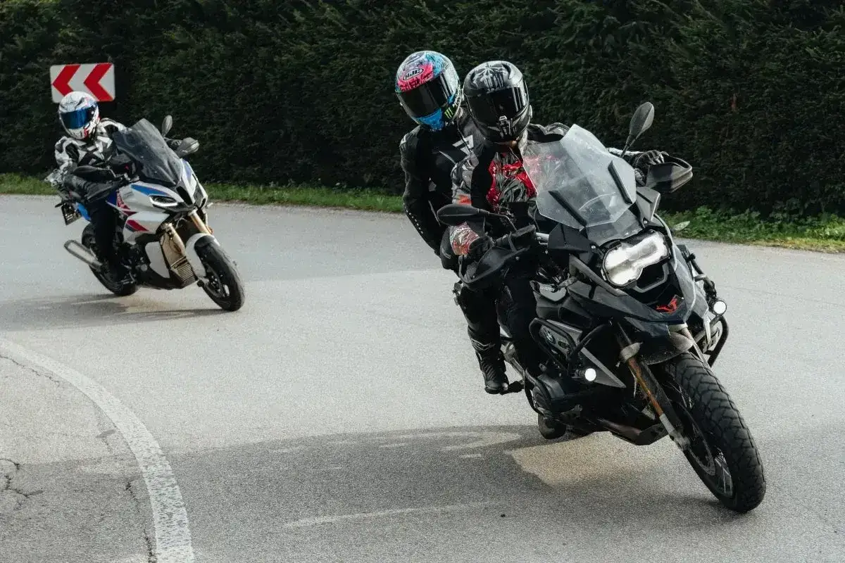 Trzech motocyklistów na zakręcie. Jeden z nich, na czarnym motocyklu, z pasażerem, wykonuje płynny międzygaz, pochylając się w zakręcie.