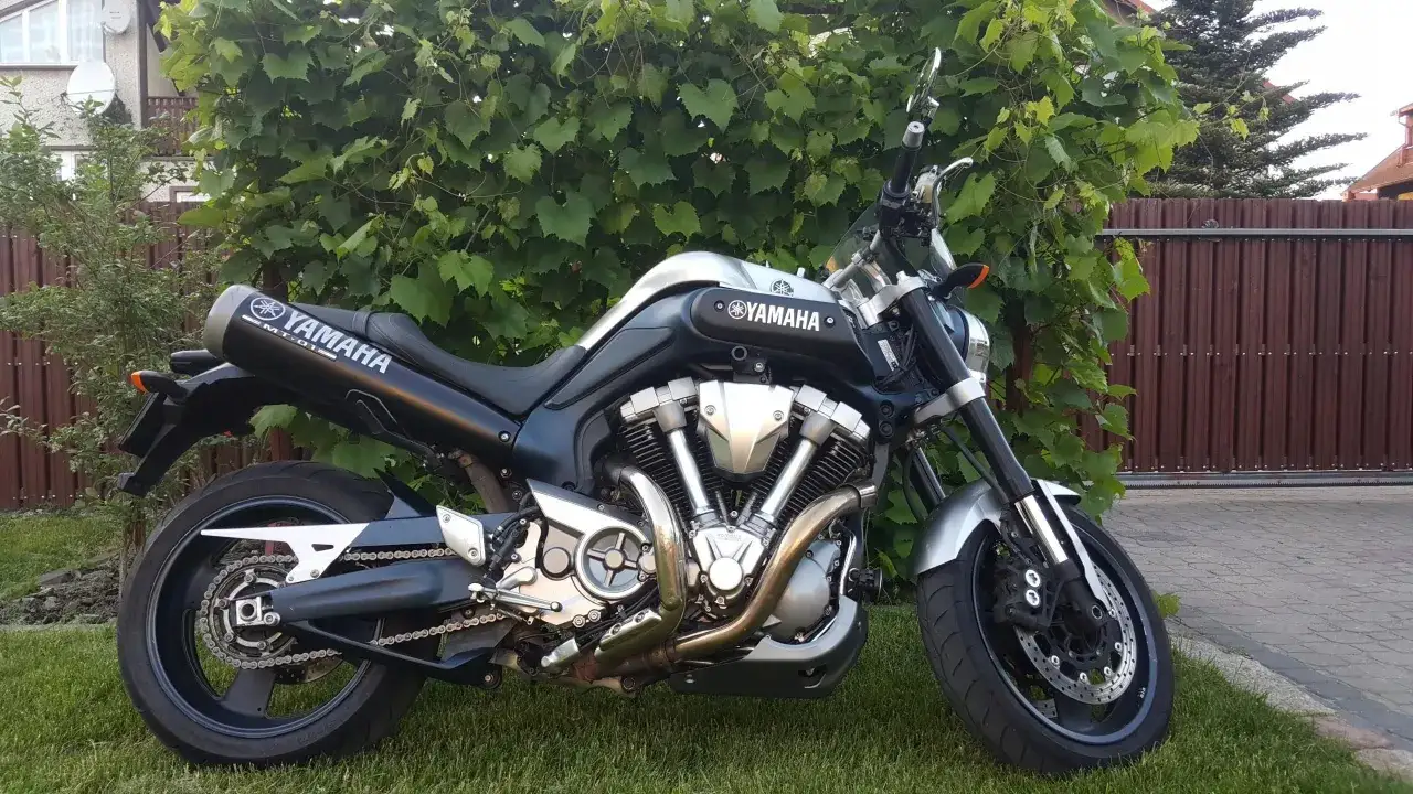 Potężny Yamaha MT-01 z charakterystycznym silnikiem V-twin, zaparkowany na trawie przed zielonym żywopłotem.