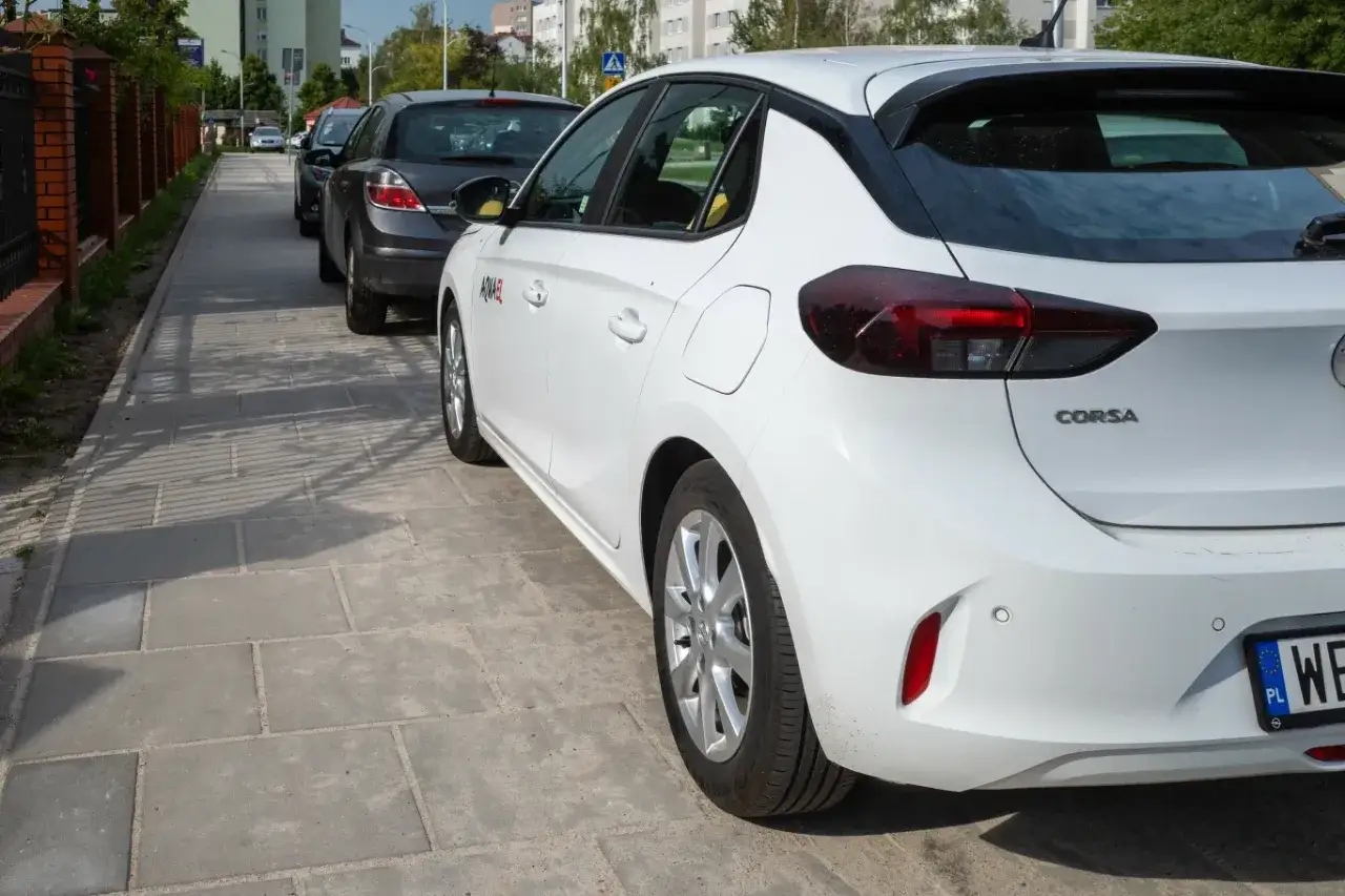 Biały Opel Corsa i inne samochody stoją na chodniku. Czy można parkować na chodniku? To pytanie zadaje sobie wielu kierowców.