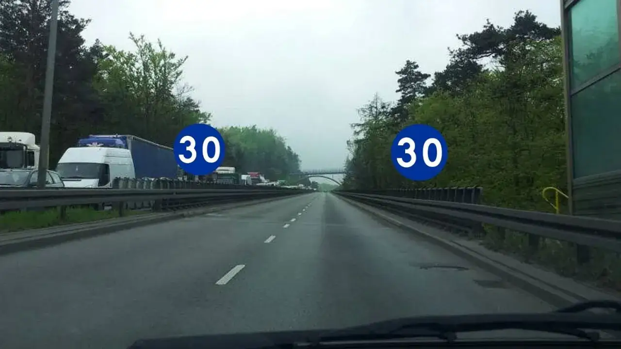 Na autostradzie znaki wskazują minimalna prędkość 30 km/h. Widać ruch drogowy i drzewa.