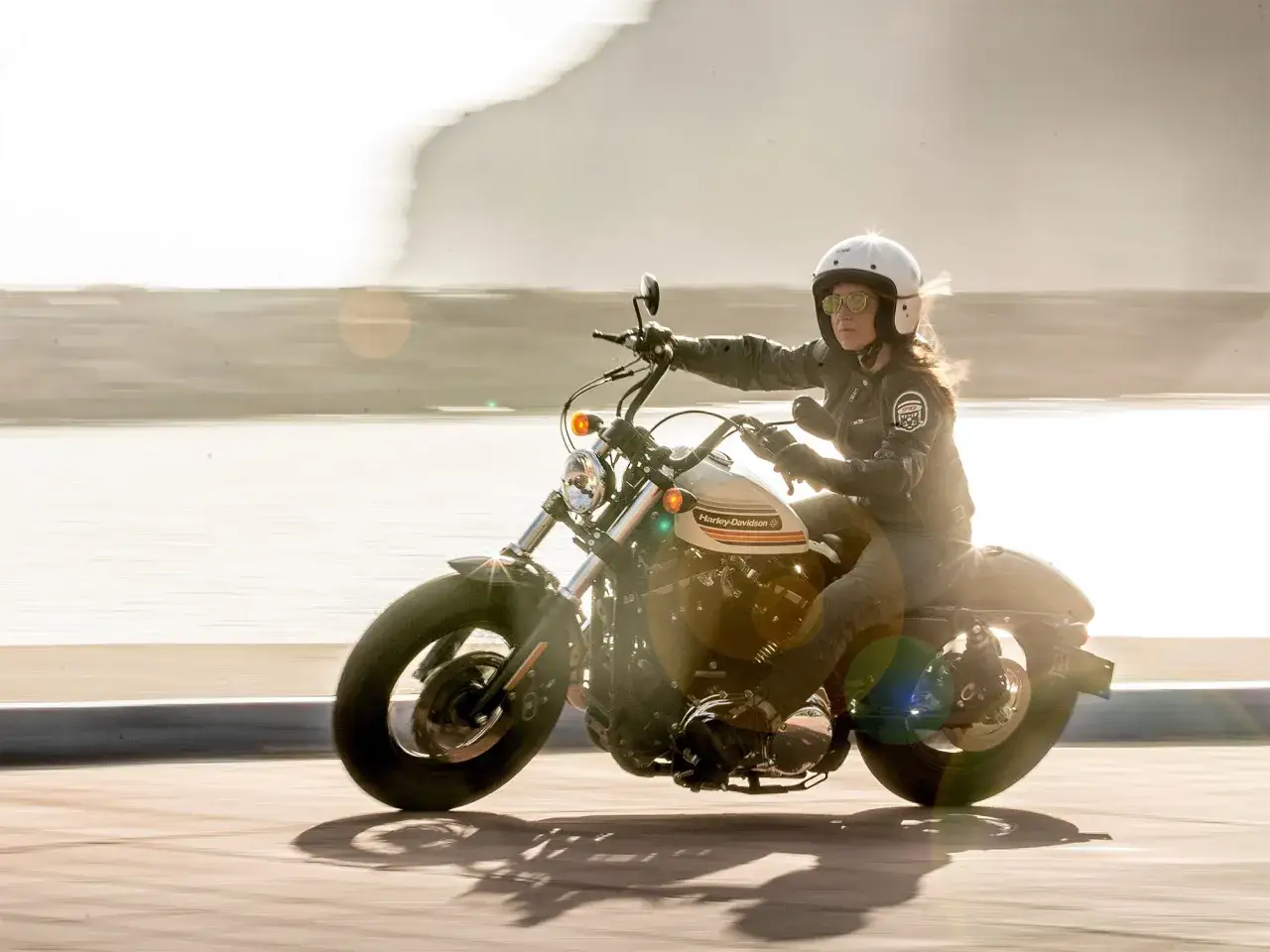 Kobieta w kasku i okularach jedzie na motocyklu typu cruiser w słoneczny dzień.