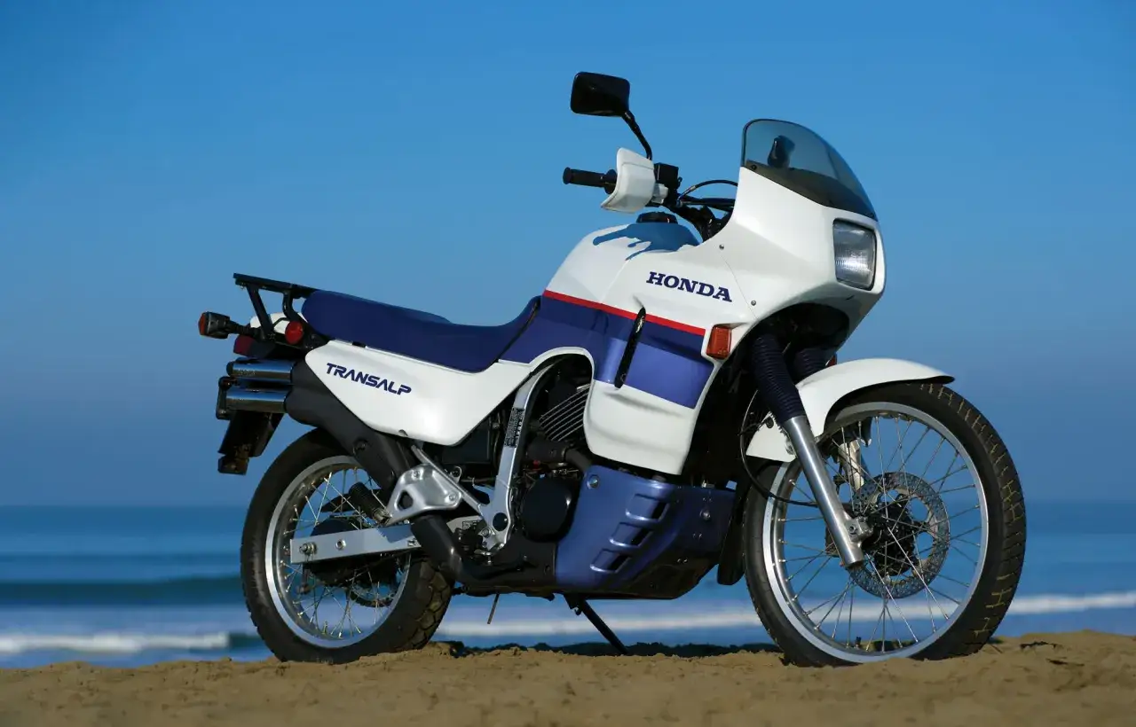 Biały motocykl Honda Transalp 650 z niebieskimi i czerwonymi akcentami stoi na plaży.