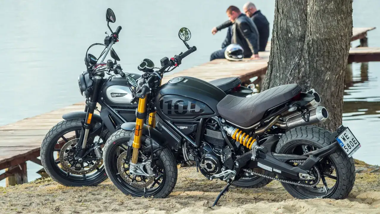 Dwa motocykle Ducati Scrambler 1100 na drewnianym pomoście nad jeziorem. Jeden czarny, drugi srebrny.