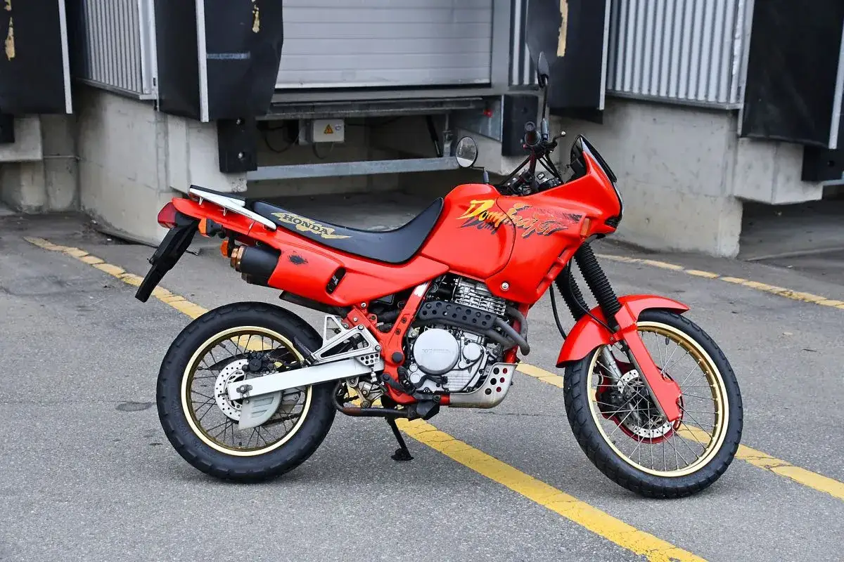 Czerwony motocykl Honda Dominator z lat 90. stoi na asfalcie. Ma złote felgi i charakterystyczne malowanie.
